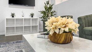 Ett modernt vardagsrum med en platt-TV som är monterad på väggen, ett vitt avlastningsbord med dekorativa föremål, en trävas med blommor och en grön soffa.