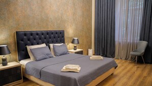 Un lit bien fait, recouvert d’une couvre-lit grise, avec deux lampes de chevet, une chaise et une fenêtre avec des rideaux.