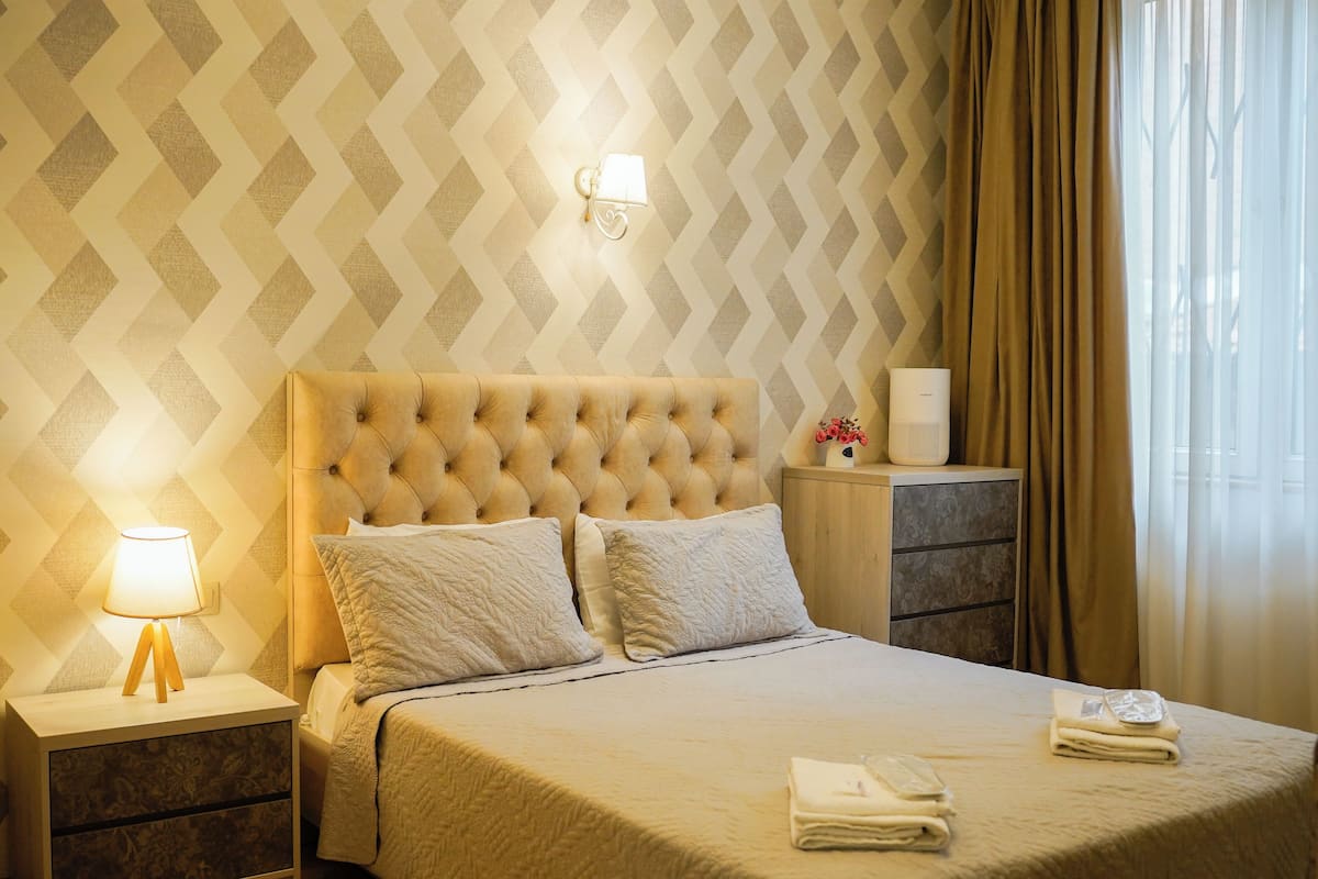 A neatly made bed with a tufted headboard, a bedside lamp, a nightstand with a floral-patterned drawer, and a bedside table with a white speaker and a small vase with flowers.