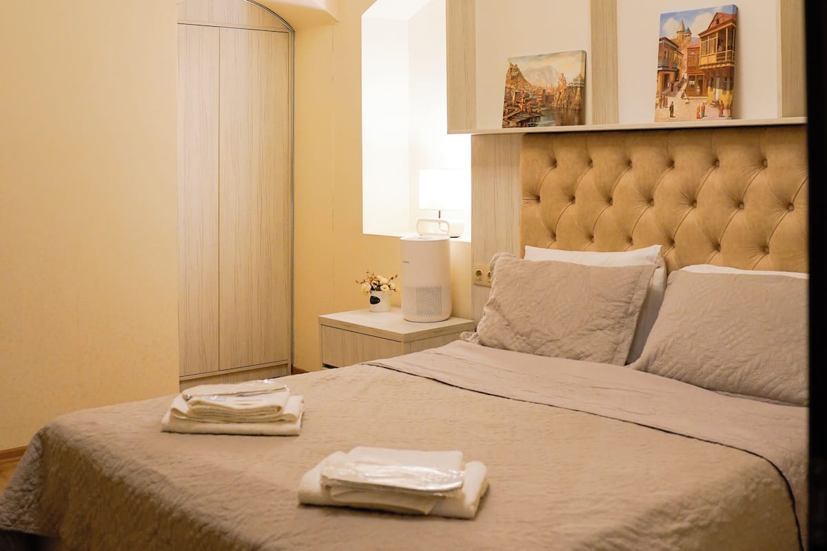 A neatly made bed with a headboard, a bedside table with a lamp, and a wall with framed pictures.
