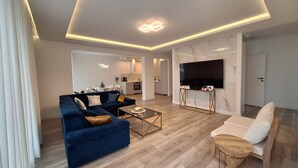 Un salon moderne avec un canapé bleu, un fauteuil blanc et une table basse. Un téléviseur est fixé au mur, et on aperçoit une cuisine au fond.