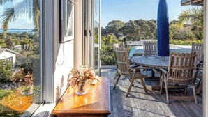 Eine Holzterrasse mit Tisch und Stühlen, eine Glastür, die zu einem Blick auf den Ozean führt, und ein blauer Sonnenschirm.