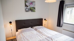 A bedroom with a dark headboard, two bedside lamps, a bedside table, a window with curtains, and a wall-mounted picture of flowers.