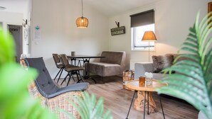 A cozy living room with a wicker sofa, a dining table with chairs, a cushioned armchair, a wooden coffee table, and a hanging light fixture.