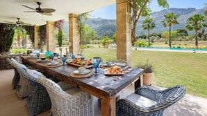 An outdoor dining area with a long wooden table, wicker chairs, and a view of mountains and a swimming pool. - Deluxe Villa Els Gorgs (Pollensa / Pollença)