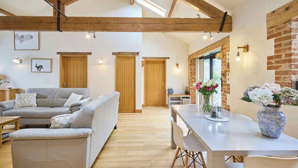 A spacious living area with a high ceiling, exposed wooden beams, a grey sofa, a dining table with chairs, and a brick wall.