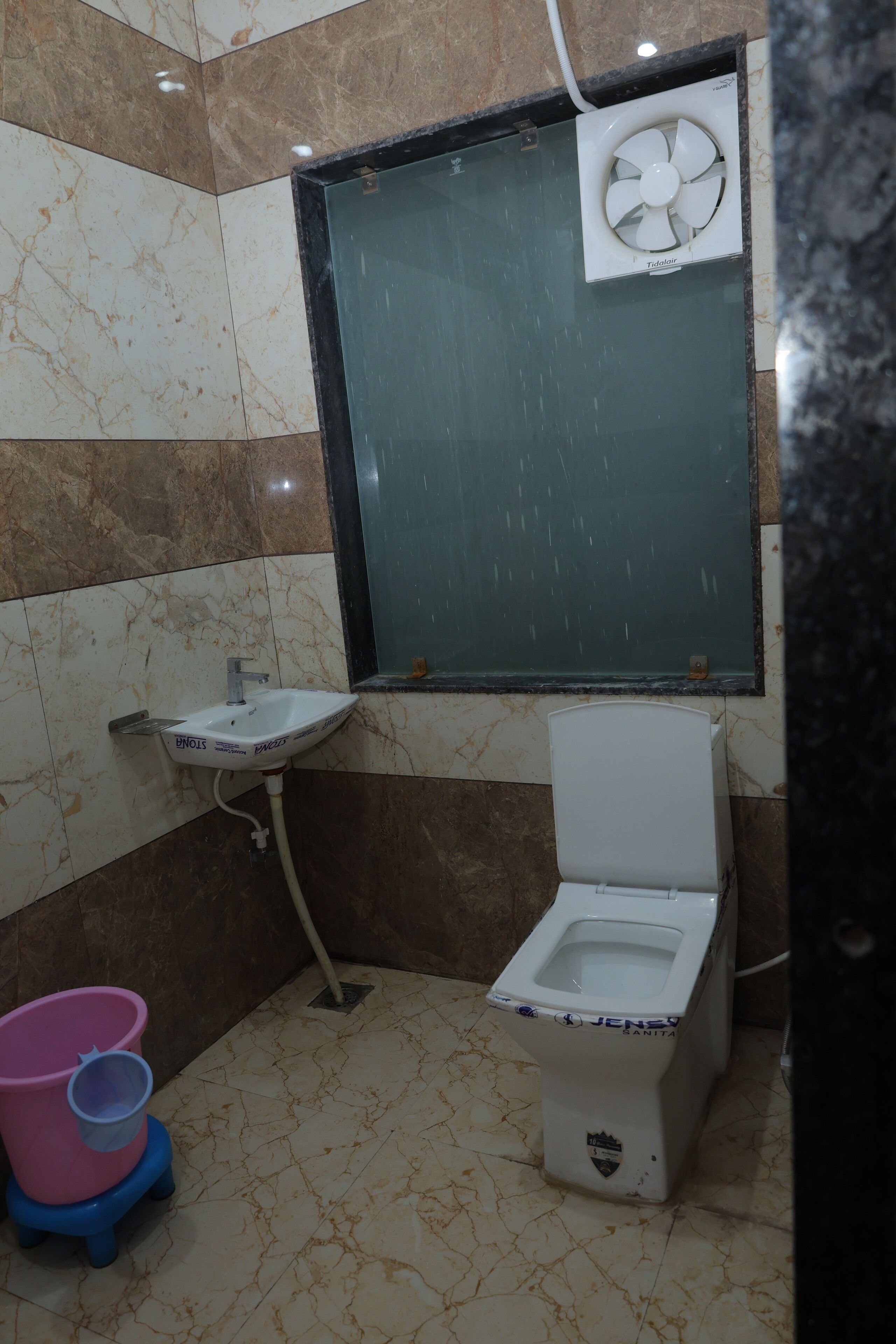 Een badkamer met een wit toilet, een wastafel op voet, een ventilator aan de muur en een roze en blauwe emmer.