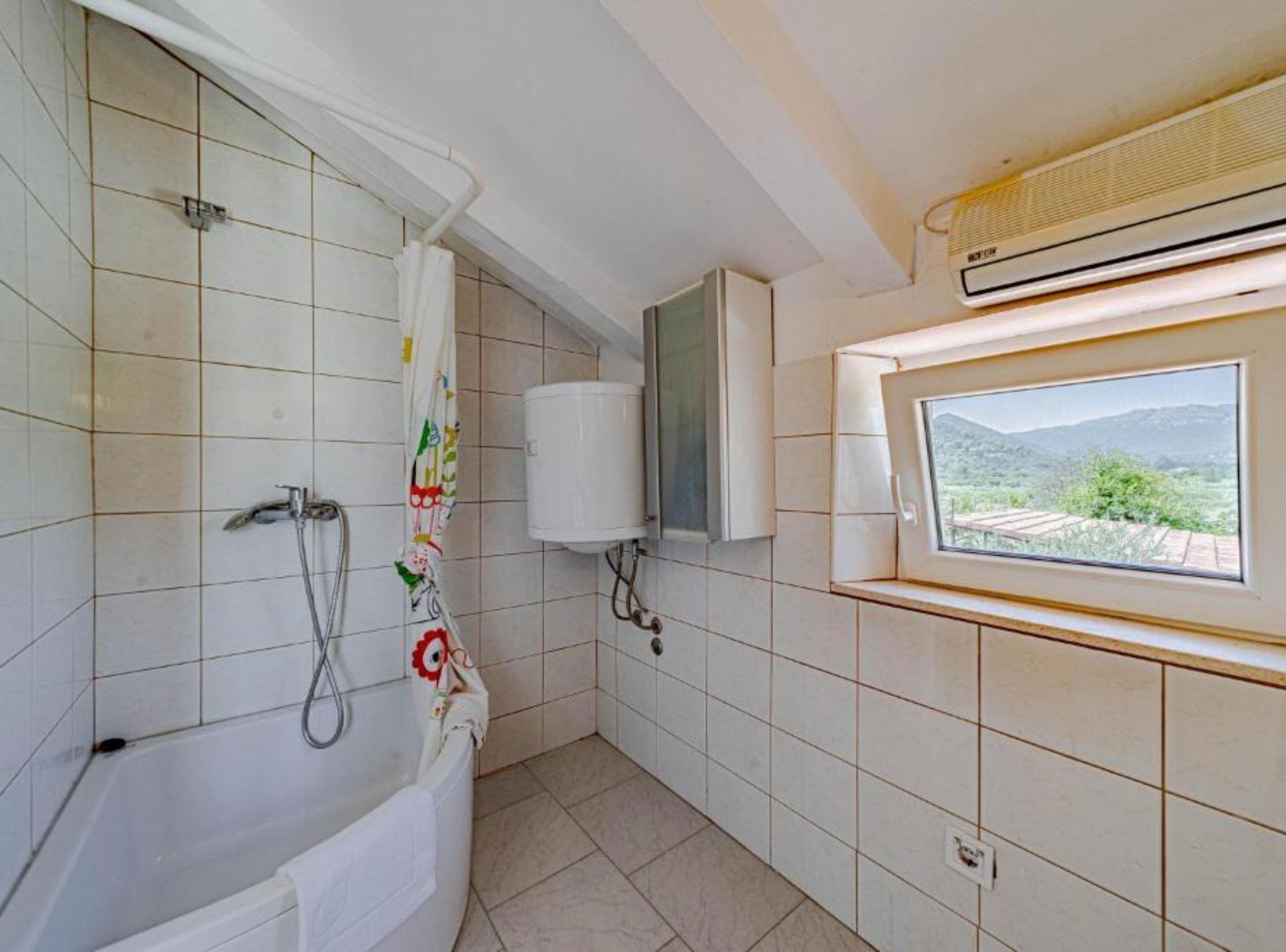 A bathroom with a bathtub, a shower, a window with a view, and a white air conditioning unit.
