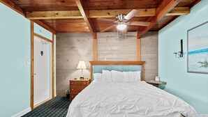 Un dormitorio con techo de madera, ventilador de techo, cama con cubrecama blanco, mesita de noche con lámpara y un cuadro en la pared.