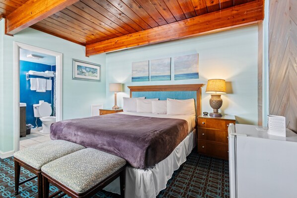 A hotel room with a wooden ceiling, a bed with a purple blanket, two bedside tables with lamps, a wooden dresser, and a patterned floor. - Oceanfront Inn Room #9 - Inlet View - King Bed (Fort Pierce)