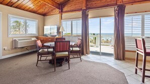 Quarto com teto de madeira, área de jantar com cadeiras e mesa, janela grande com persianas e vista para o oceano.