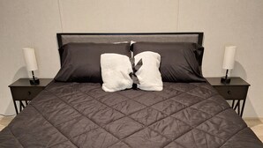 Ein großes Bett mit dunkler, gesteppter Bettdecke, zwei Nachttische auf jeder Seite und eine Wandlampe auf jedem Nachttisch.