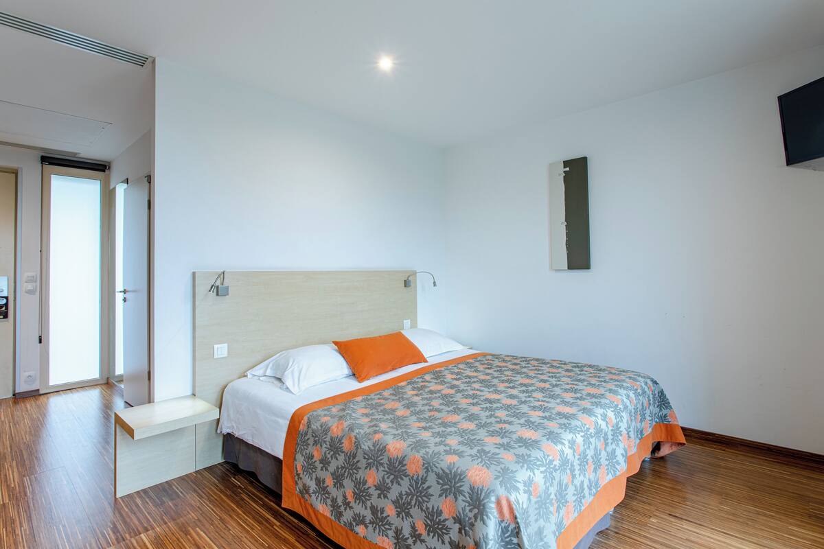 Une chambre à coucher avec un lit, un banc, un téléviseur fixé au mur et une porte.