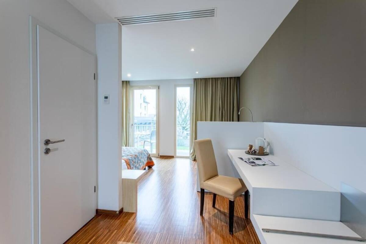 Une chambre d’hôtel moderne avec un plancher en bois, un lit recouvert d’une couverture colorée, un bureau avec une chaise et une vue sur l’extérieur.