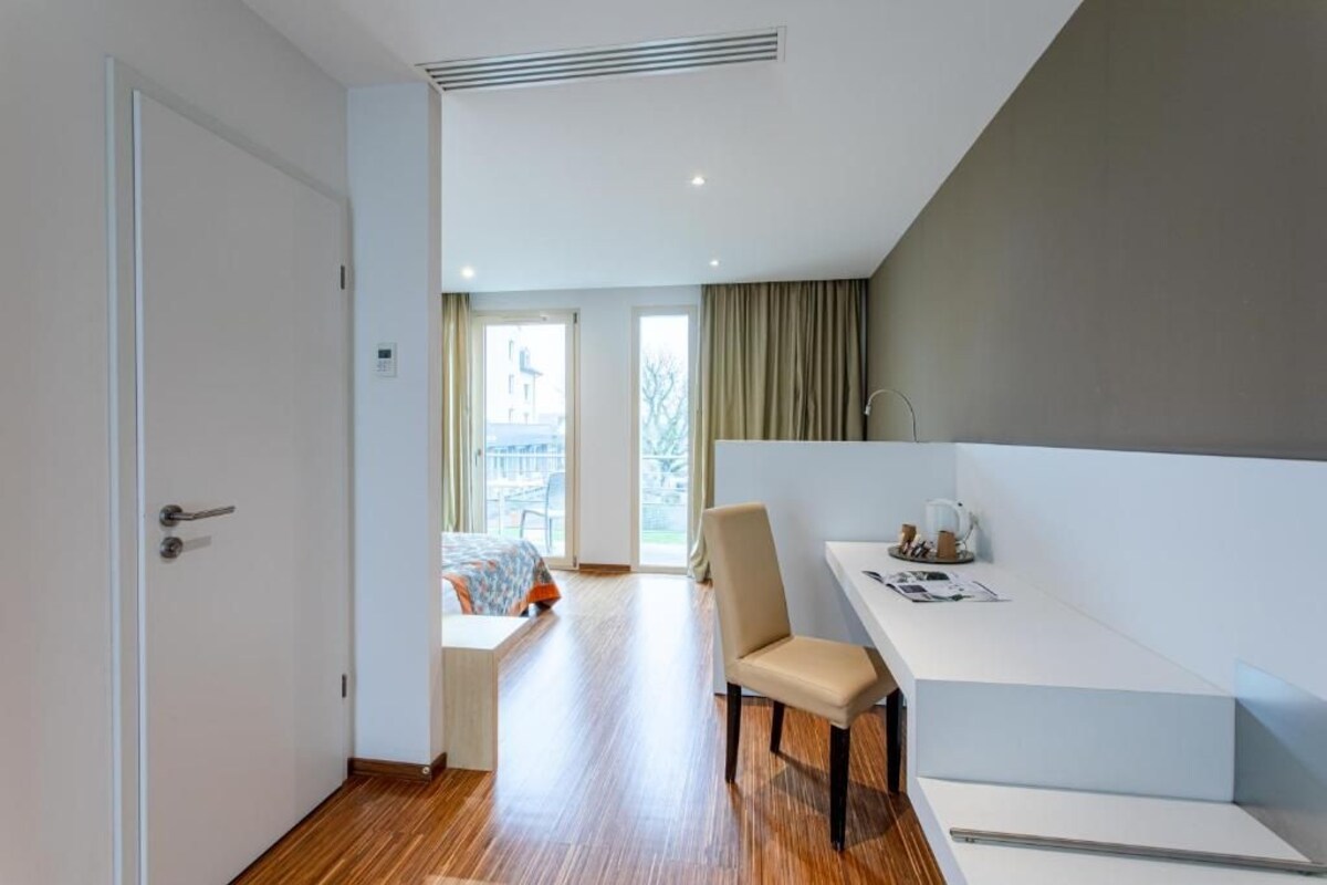 Une chambre d’hôtel moderne avec un plancher en bois, un lit recouvert d’une couverture colorée, un bureau avec une chaise et une vue sur l’extérieur.
