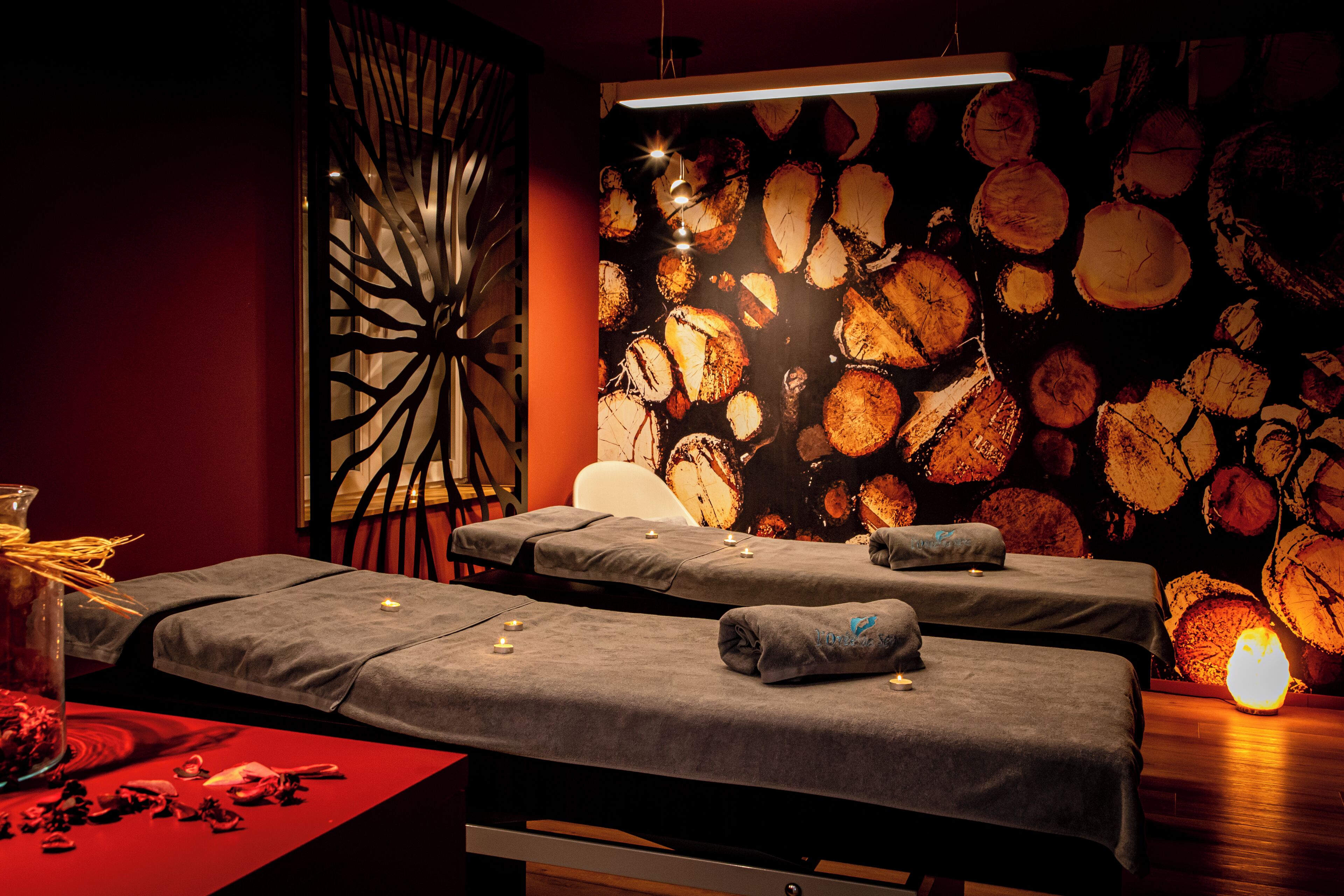 Een spa-ruimte met twee massagetafels, een decoratieve muur met cirkelvormige patronen, en een glazen vaas met gedroogde bloemen op een rode tafel.