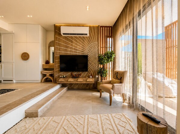 A modern living room with a flat-screen TV, a wooden accent wall, a large window with sheer curtains, and a seating area with a sofa and armchair.