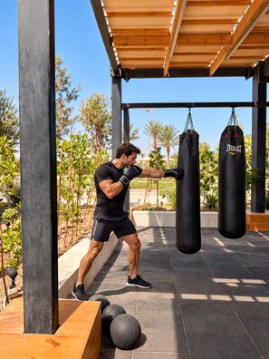 A person training with a punching bag in an outdoor gym area with a wooden pergola overhead.