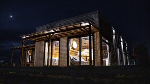 A modern, well-lit cabin with large windows, a wooden exterior, and a visible interior with a bed and a round light fixture.