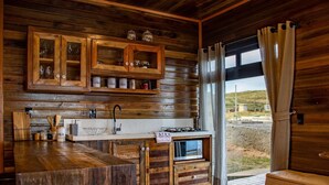 Interior rústico de uma cabana de madeira, com área de cozinha, armários de madeira, pia e vista para o exterior.