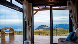 Quarto com grande janela com vista para as montanhas, deck de madeira, sofá com almofadas e tapete estampado.