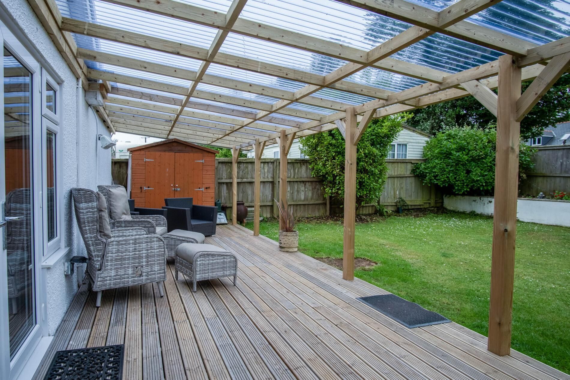Eine überdachte Terrasse mit Holzdeck, Gartenmöbeln und einem Holzschuppen im Hintergrund.
