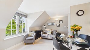 A modern loft bedroom with a sloped ceiling, a dining area with a glass table, and a view of greenery through a large window. - 29 Rhodewood House - 2 Bedroom - Saundersfoot (Saundersfoot)