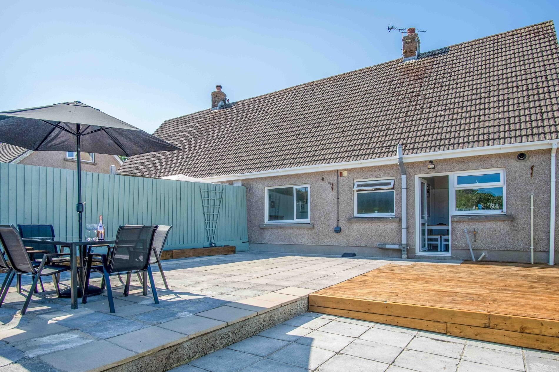 A residential property with a tiled roof, a wooden deck, a patio with a table and chairs, and a blue fence.