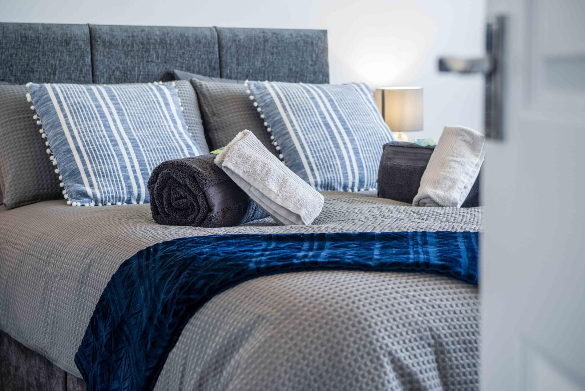 Un letto rifatto con ordine, con una testiera grigia, cuscini a righe e una coperta blu.