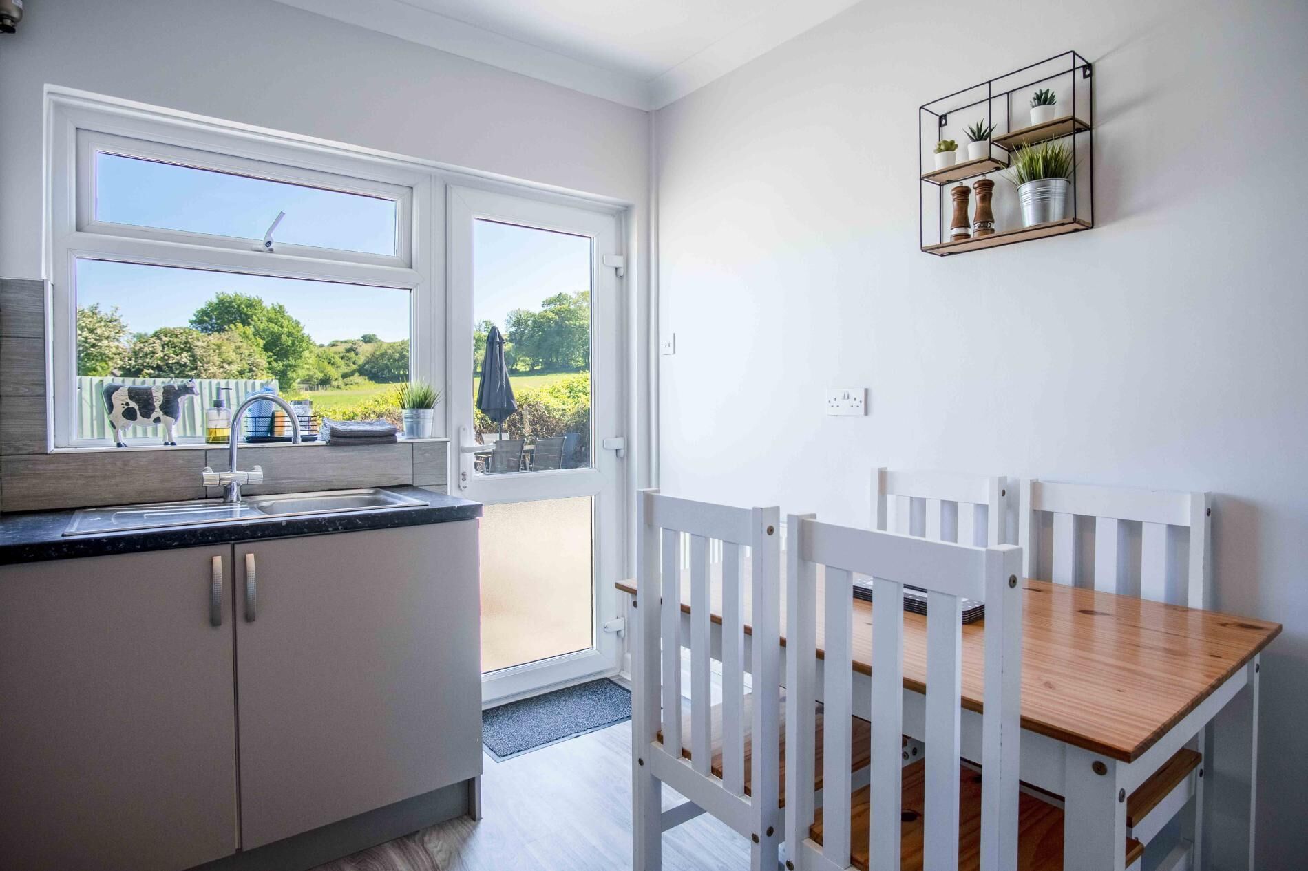 A kitchen with a window, a dining area with a white table and chairs, and a wall-mounted shelf with plants.