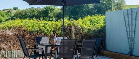 Een terras buiten met een tafel en stoelen, een grote parasol, en een uitzicht op een haag en bomen op de achtergrond.