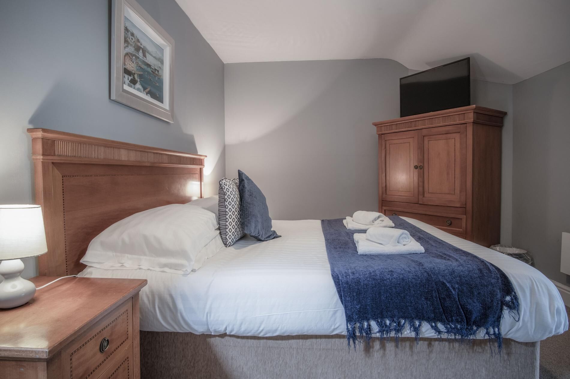 A neatly made bed with a patterned pillow, a blue throw blanket, and folded towels. There is a wooden nightstand with a lamp and a framed picture on the wall above the bed.