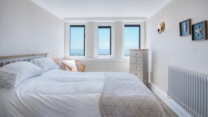 A bedroom with a large bed, a dresser, and a view of the sea through three windows. - Waterfront 1 - 1 Bedroom Apartment - Wisemans Bridge (Saundersfoot)