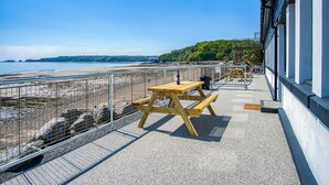 A balcony with picnic tables overlooking a beach, a railing, and a building with large windows. - Waterfront 1 - 1 Bedroom Apartment - Wisemans Bridge (Saundersfoot)