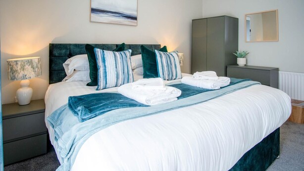 A neatly made bed with white linens, a teal throw blanket, and matching pillows. There is a bedside lamp, a small potted plant, and a mirror on the nightstand. - No 7 Moorfield Road - 3 Bed Townhouse - Narberth (Narberth)