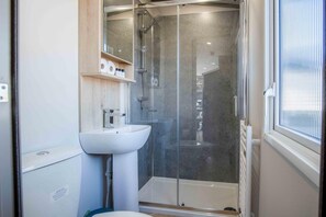 Ein modernes Badezimmer mit einer in Glas abgeschlossenen Dusche, einem weißen Waschbecken und einem Holzregal darüber.