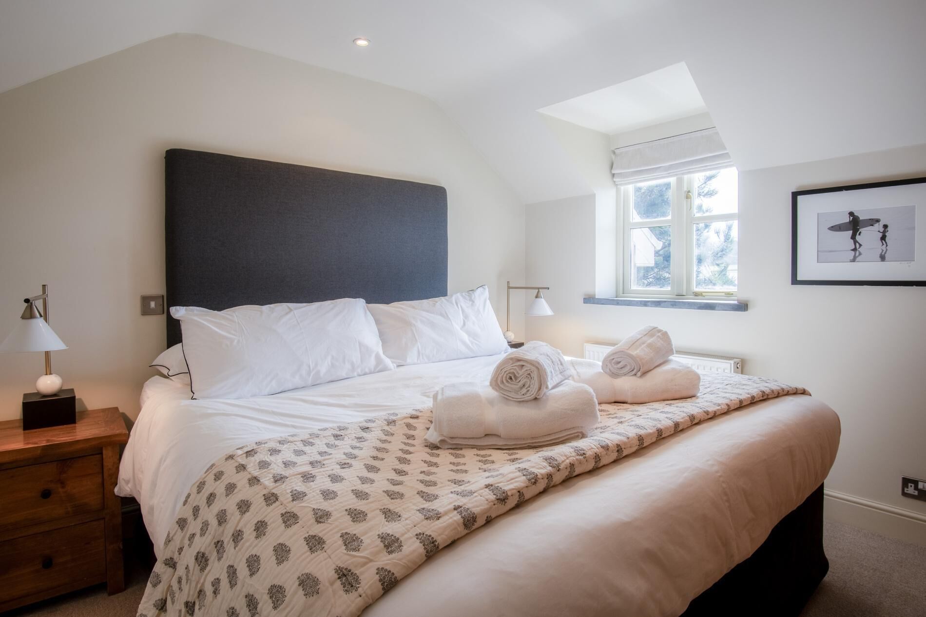 A neatly made bed with white linens and rolled towels, a bedside table with a lamp, and a window with a view of trees.