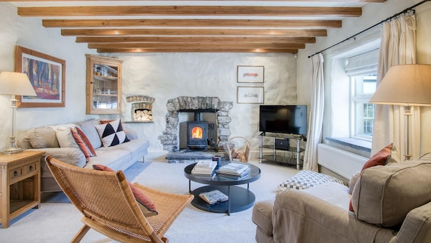A cozy living room with a stone fireplace, wooden beams, a sofa, a wicker chair, and a TV. - Fern Cottage - 2 Bedroom Cottage - St. Florence (Tenby)