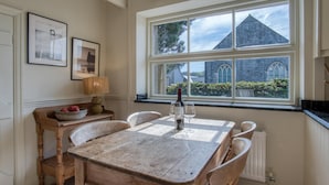 A dining area with a wooden table and chairs, a window with a view of a building, and a lamp on a side table. - Fern Cottage - 2 Bedroom Cottage - St. Florence (Tenby)