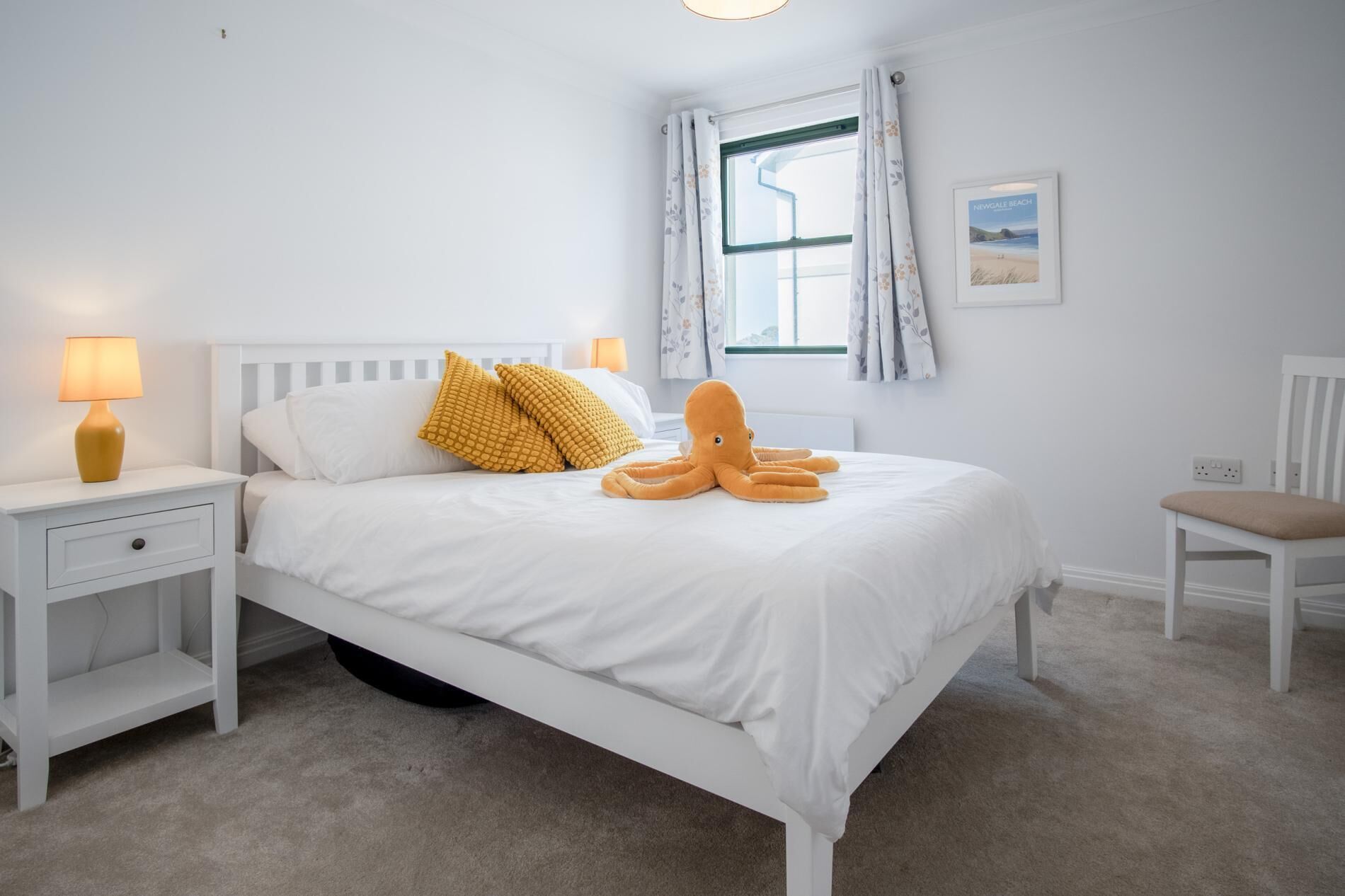 Een slaapkamer met een bed, een nachtkastje, een stoel, een raam met gordijnen en een aan de muur hangend schilderij.