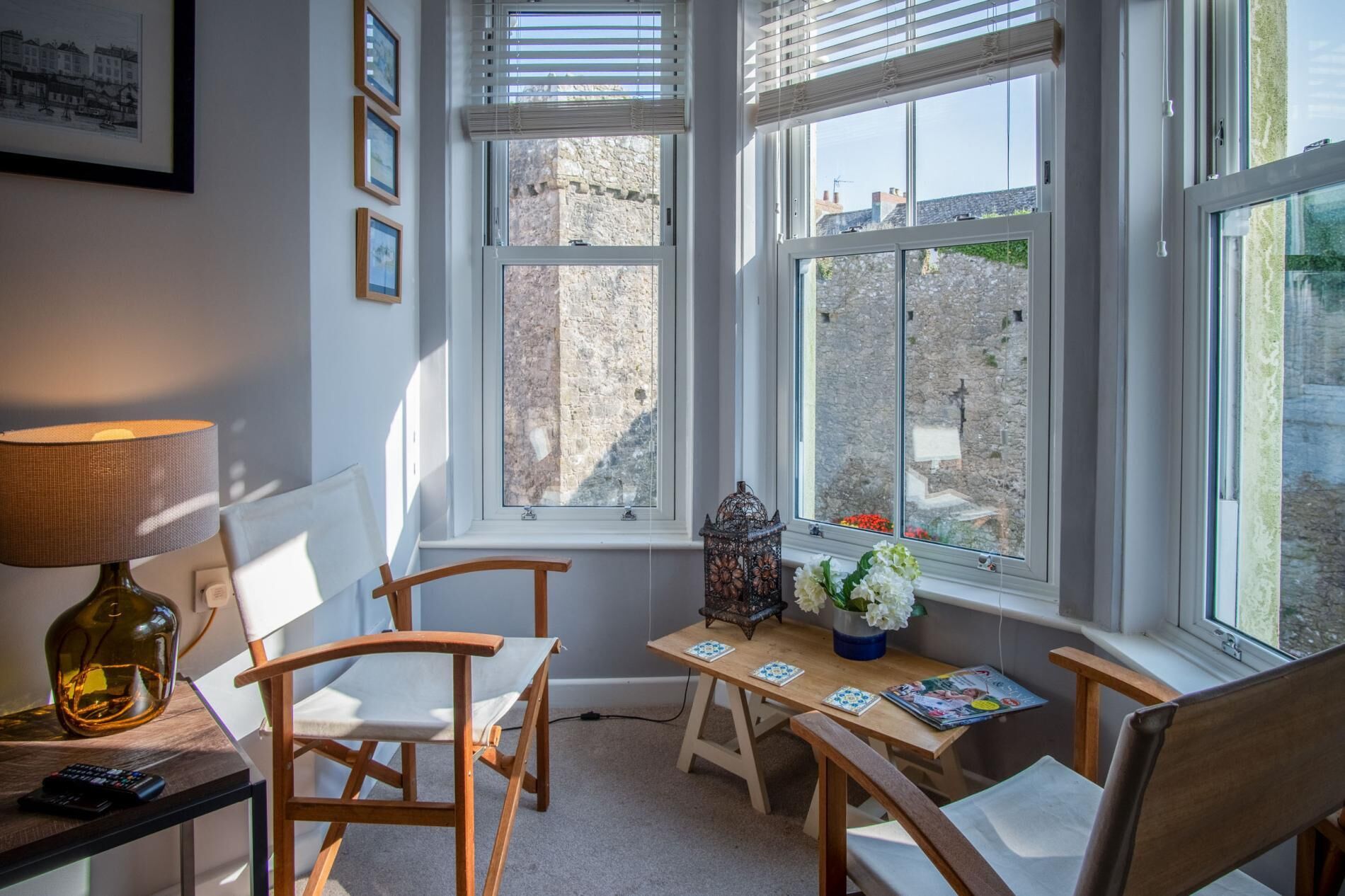 A cozy room with a wooden table and chairs, a lamp, and a view of stone buildings through large windows.