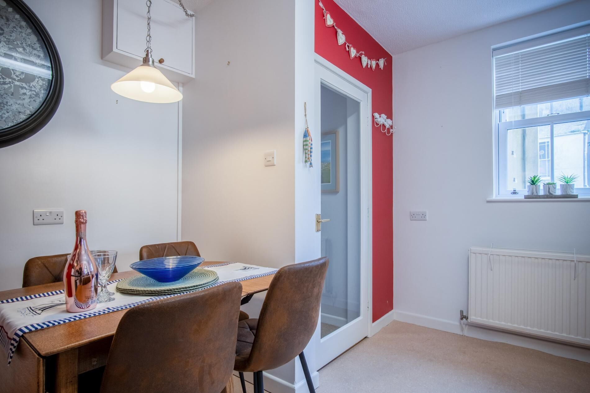 A dining area with a table set for two, a bottle of champagne, and a blue bowl. There is a red wall with a heart garland, a window with blinds, and a radiator.