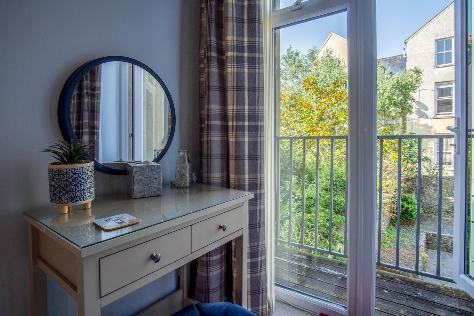 A bedroom with a glass dresser, a round mirror, a potted plant, and a view of a balcony with a railing and greenery.