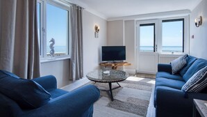Ett vardagsrum med en TV, två soffor, ett runt soffbord och stora fönster med utsikt över havet.