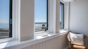 Ett rum med utsikt över stranden genom stora fönster, en flätad fåtölj med en kudde och en liten hylla med dekorativa föremål.