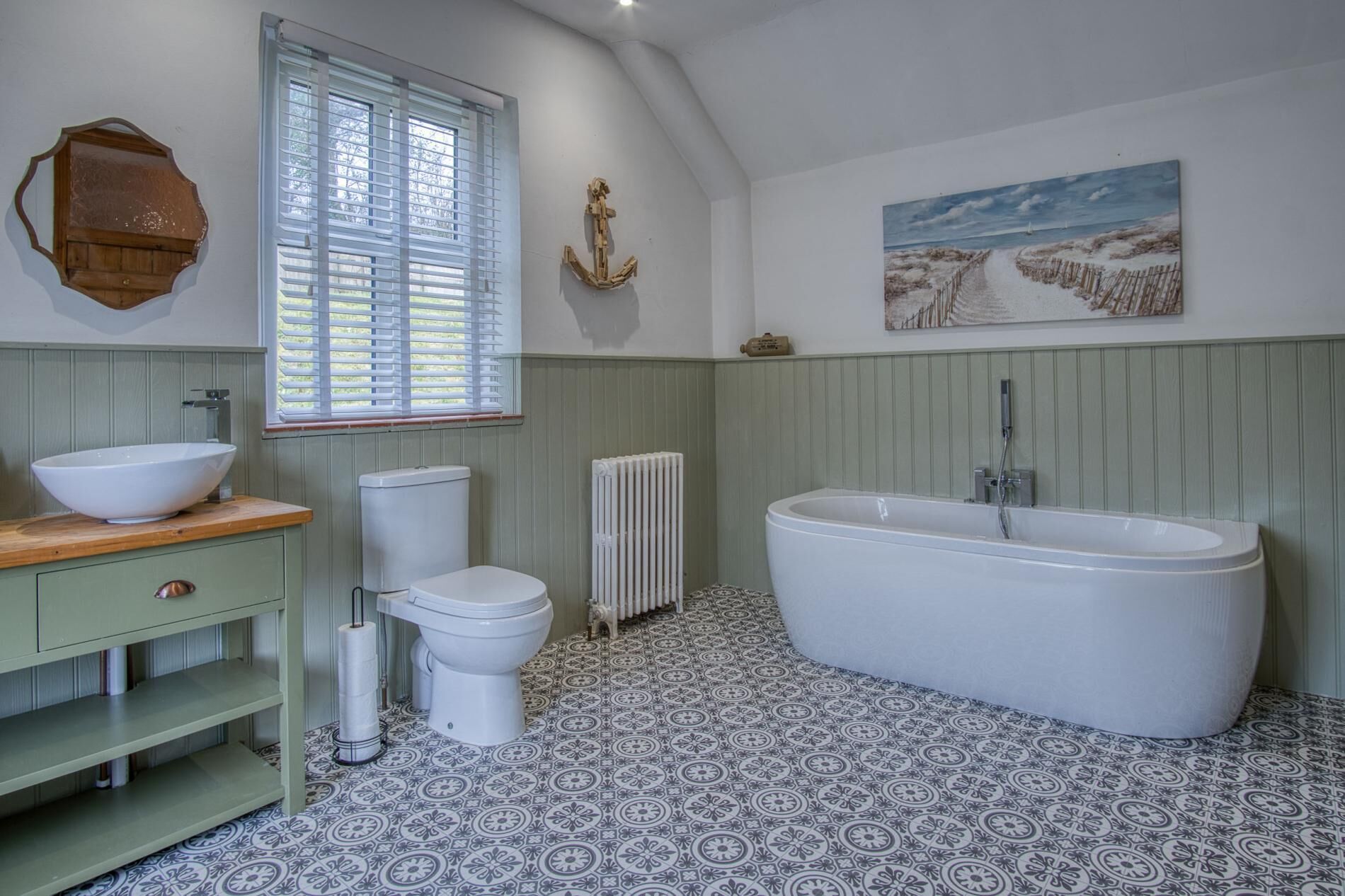A bathroom with a freestanding bathtub, a toilet, a sink with a wooden countertop, patterned floor tiles, a window with blinds, and a wall-mounted painting.