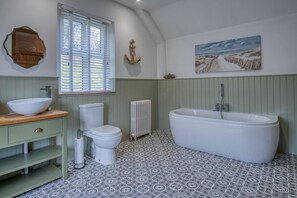 A bathroom with a freestanding bathtub, a toilet, a sink with a wooden countertop, patterned floor tiles, a window with blinds, and a wall-mounted painting. - Woodcocks House - 3 Bedroom Holiday Home - Kilgetty (Kilgetty)