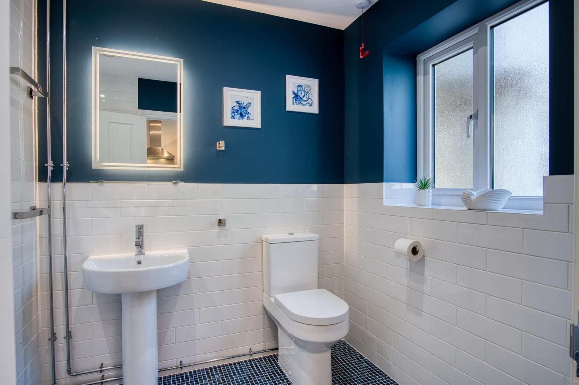 Ein Badezimmer mit weißem Waschbecken, Toilette und Spiegel. An der Wand hängen zwei Bilder in Rahmen.