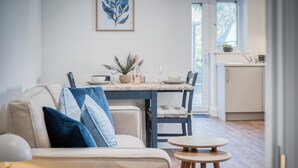 Una sala de estar moderna con un sofá, una mesa de comedor dispuesta con platos, vasos y un florero. Se ve una cocina al fondo con armarios y una ventana.