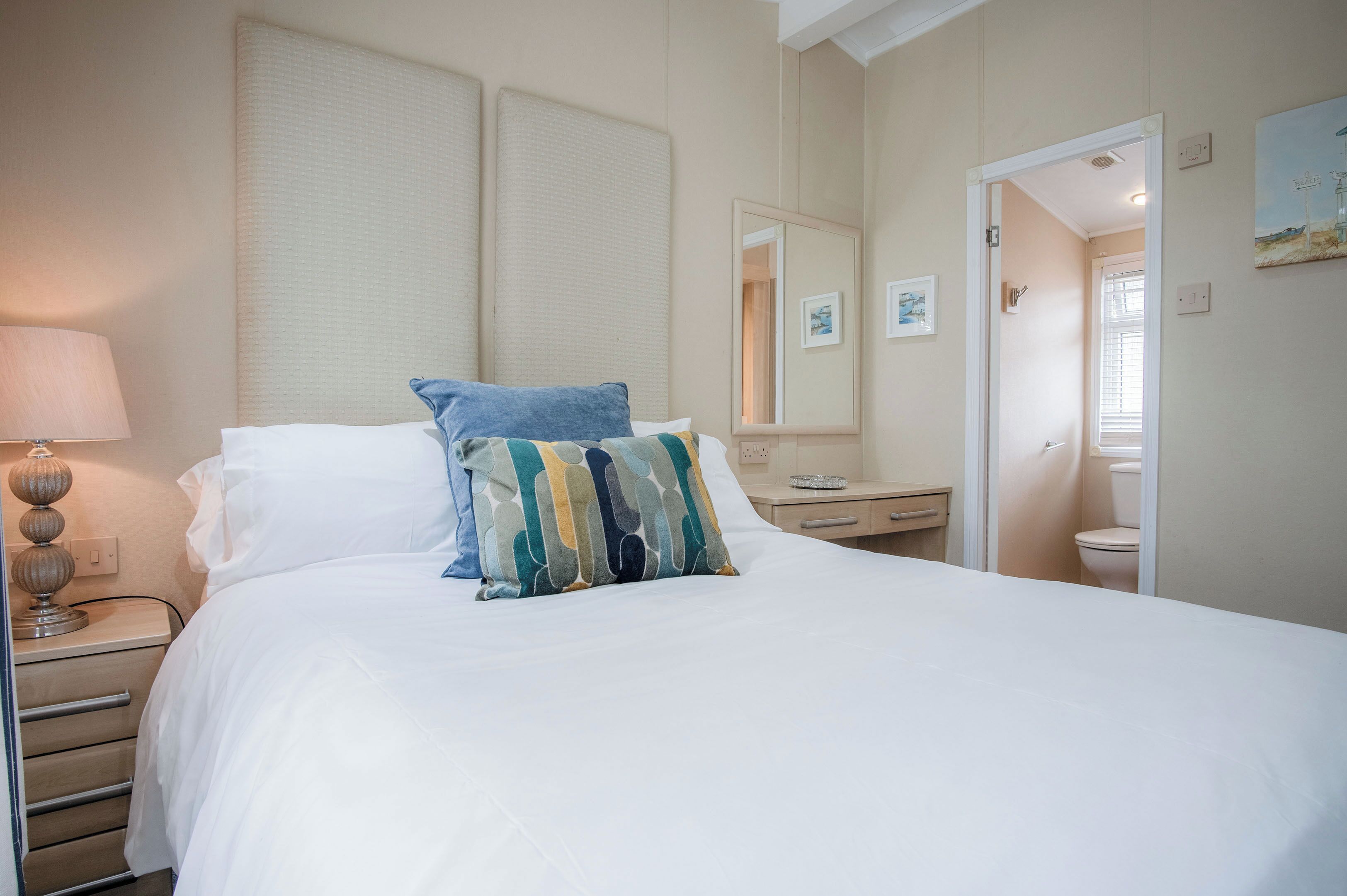 A hotel room with a large bed, a bedside table with a lamp, a small bedside table with a tray, a mirror, and a door leading to a bathroom.